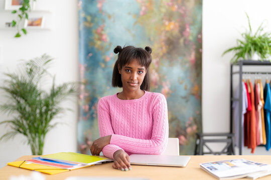 A young Black woman with space buns sits at a desk with a laptop and folders, looking at the camera. She wears a pink sweater in a creative workspace.