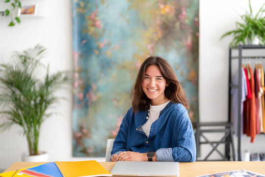A cheerful young woman with long brown hair smiles at the camera while sitting at a wooden desk. A laptop and colorful folders are in front of her in a bright, modern workspace.