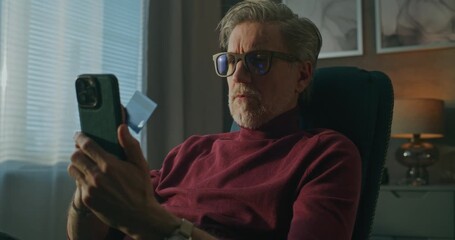 Aged Man in Glasses Uses Smartphone and Credit Card Complete Online Transaction While Relaxing in Hotel Chair. Concept Secure Online Shopping, Convenient Digital Payments, Ecommerce While Traveling. - Powered by Adobe