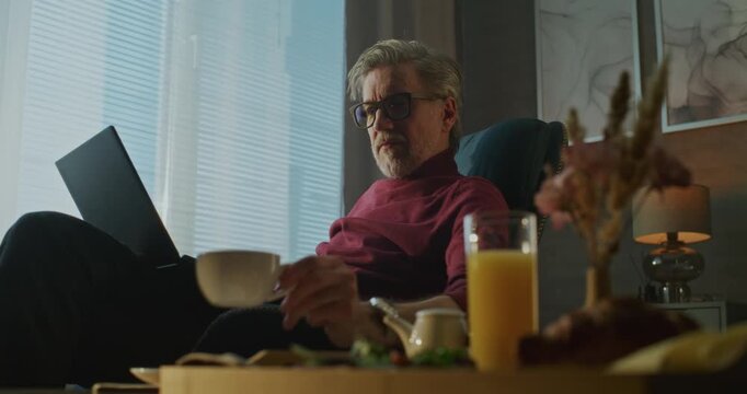 Gray Haired Man Sits Relaxed in Hotel Chair, Sipping Coffee and Typing on Laptop in Morning Light and Cozy Atmosphere. Concept of Remote Work, Business Travel, and Digital Flexibility. Handheld.