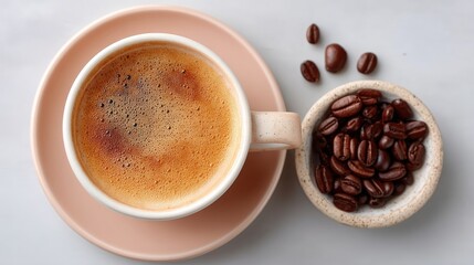 Fototapeta premium Flat lay overhead view of a ceramic cup filled with frothy coffee beside a small bowl of roasted coffee beans on a light gray marble surface
