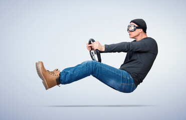 Man wearing pilot goggles floating above the ground in a driver’s pose, holding a car steering wheel on a light background. Concept of imagination, speed, travel and freedom.