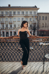 Stylish woman in black jumpsuit posing by balcony railing
