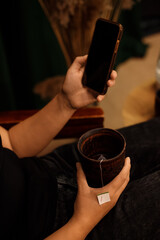 A person relaxes with a cup of tea, a phone, and an electric kettle in a cozy home atmosphere