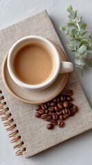Overhead view of a white ceramic cup filled with frothy coffee and scattered roasted coffee beans on a sparkling patterned notebook with a sprig of eucalyptus on a gray background.