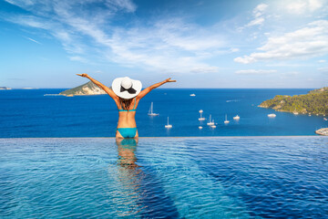 A happy woman sitting at the edge of an infinity pool and enjoying the view of the mediterranean sea during her summer vacations