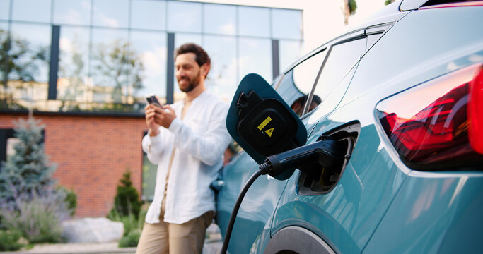 Electric car charging at portable station on city street, man using smartphone app in background, urban buildings, eco-friendly transportation concept.