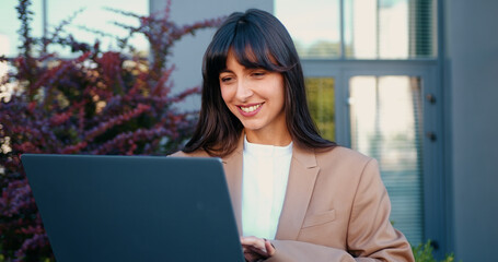Close-up of a young professional woman working on a laptop outdoors, smiling confidently as she types and reviews information on the screen.