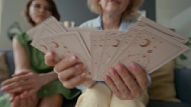 Close-up low angle shot of middle-aged Caucasian woman shuffling tarot cards, while her friend waiting, sitting on couch in living room