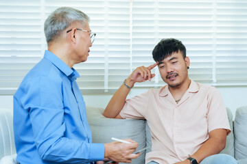 Obraz premium Asian adult man pointing to head during counseling with senior psychiatrist, discussing emotional challenges and receiving professional guidance for stress management and psychological healing