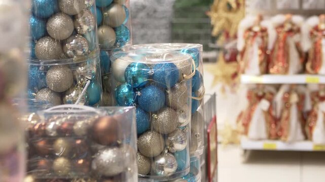 Blue silver ornament packs beside nativity figures, store aisle scene with packaged baubles and figurines in background, assistant restocking seasonal inventory, warm cozy lighting