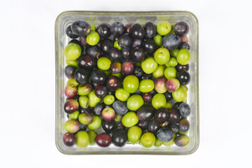 An overhead shot of a square glass container filled entirely with an assortment of fresh green, black, and purplish olives, centered on a white background