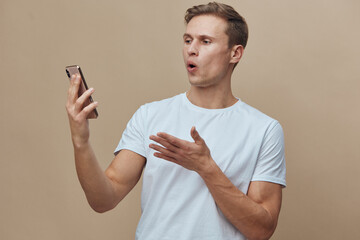 surprised young caucasian man talking on phone in white t-shirt focused on screen using mobile device isolated on beige background digital communication concept