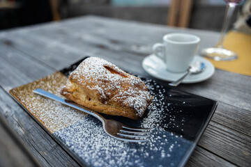 fetta di strudel di mele su un piatto nero, vicino ad una tazza di caffè, su un tavolo di legno, a pranzo
