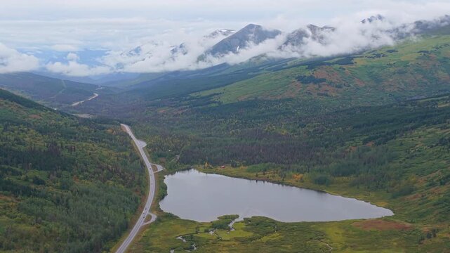 Aerial view of a winding road alongside a serene lake, surrounded by lush green forests and mountains shrouded in mist, Seward, Alaska, United States.