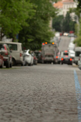 This scene features a charming cobblestone street with colorful vehicles, lush trees, and a nearby service van, creating a vibrant and welcoming urban atmosphere for passersby