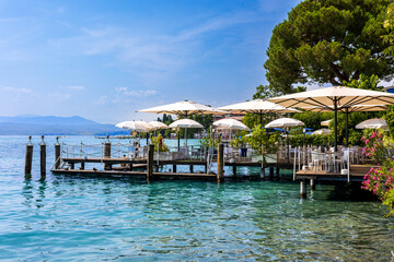 Relaxing by the sparkling waters of Sirmione at Lake Garda, meals on a wooden dock at Sirmione, surrounded by the blue waters of Lake Garda. Bright umbrellas provide shade on this sunny day.