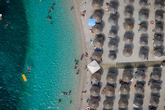 Aerial view of sun-drenched beach where azure waters meet the shore, dotted with orderly rows of umbrellas offering respite from the summer heat, Ksamil, Vlor, Albania.