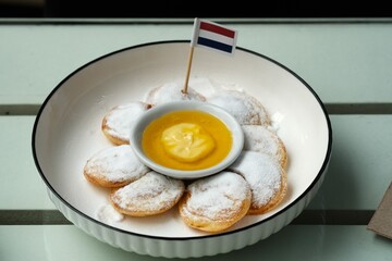 A plate of traditional Dutch mini pancakes (poffertjes) dusted with powdered sugar, served with a custard dip and a Dutch flag topper.	
