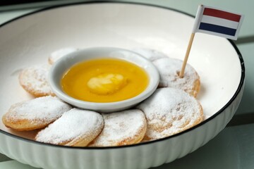 A plate of traditional Dutch mini pancakes (poffertjes) dusted with powdered sugar, served with a custard dip and a Dutch flag topper.
