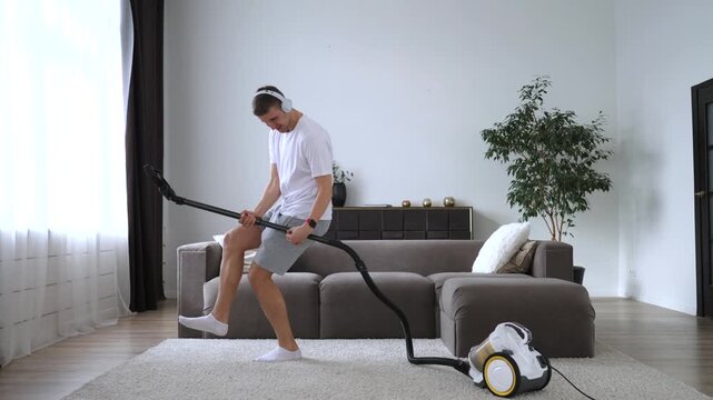 Cheerful young man wearing headphones having fun and dancing in his living room, pretending to play rock music on a vacuum cleaner hose instead of an electric guitar while doing chores
