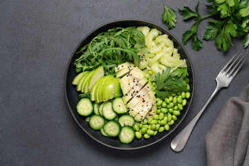 Healthy green salad with avocado, celery, apple, arugula, edamame beans, cucumber, and parsley in black bowl on dark background, top view
