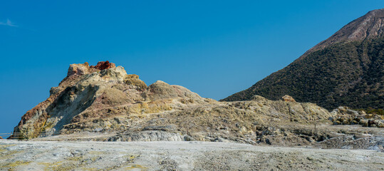 Volcanic landscape with sulfurous ground, geothermal vents, and exposed rock against a clear blue sky.