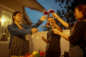 An Asian woman and friends toast with wine during a cozy backyard BBQ. Grilling skewers over charcoal under string lights, they celebrate friendship, joy,  togetherness in a festive outdoor evening