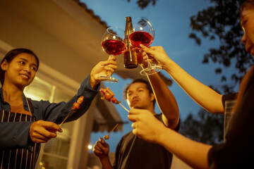 An Asian woman and friends toast with wine during a cozy backyard BBQ. Grilling skewers over charcoal under string lights, they celebrate friendship, joy,  togetherness in a festive outdoor evening