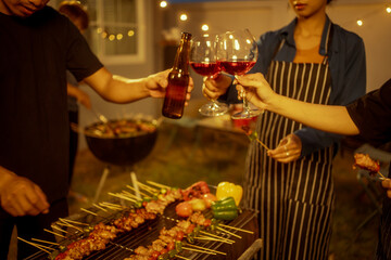 An Asian woman and friends toast with wine during a cozy backyard BBQ. Grilling skewers over charcoal under string lights, they celebrate friendship, joy,  togetherness in a festive outdoor evening