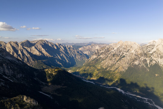 Aerial view of rugged peaks cast long shadows over the valley floor, a river snaking through the landscape below, Teth, Shkoder County, Albania.