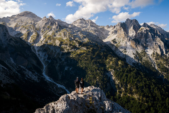 Aerial view of two figures standing atop a rocky peak amidst the rugged, verdant landscape of the Albanian Alps, bathed in soft sunlight., Teth, Shkoder County, Albania.