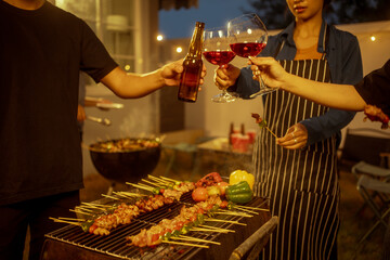 An Asian woman and friends toast with wine during a cozy backyard BBQ. Grilling skewers over charcoal under string lights, they celebrate friendship, joy,  togetherness in a festive outdoor evening