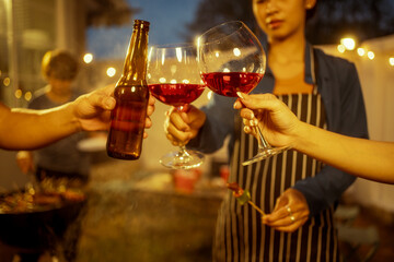 An Asian woman and friends toast with wine during a cozy backyard BBQ. Grilling skewers over charcoal under string lights, they celebrate friendship, joy,  togetherness in a festive outdoor evening