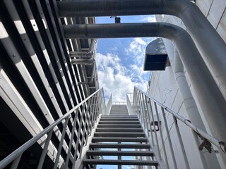 Obraz premium Low-angle view up a galvanised steel industrial staircase, flanked by large metal pipes and structures, leading towards a bright blue sky with white clouds, symbolising ascent and facility access.