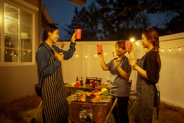An Asian woman and friends toast with wine during a cozy backyard BBQ. Grilling skewers over charcoal under string lights, they celebrate friendship, joy,  togetherness in a festive outdoor evening