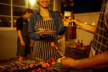 An Asian woman and friends toast with wine during a cozy backyard BBQ. Grilling skewers over charcoal under string lights, they celebrate friendship, joy,  togetherness in a festive outdoor evening