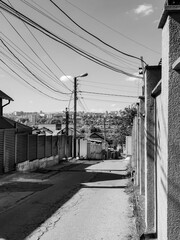 A black and white image of a narrow, winding street lined with houses and fences, leading towards a distant cityscape under a bright, cloudy sky