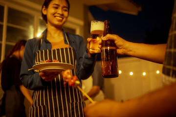 An Asian woman and friends toast with wine during a cozy backyard BBQ. Grilling skewers over charcoal under string lights, they celebrate friendship, joy,  togetherness in a festive outdoor evening