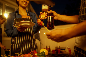 An Asian woman and friends toast with wine during a cozy backyard BBQ. Grilling skewers over charcoal under string lights, they celebrate friendship, joy,  togetherness in a festive outdoor evening