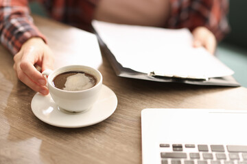 Coffee break. Woman with cup of aromatic drink and clipboard at wooden desk indoors, closeup