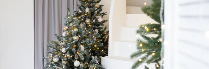 A large Christmas tree in the living room, decorated with garlands and light balloons. Bright Christmas interior with a white staircase.