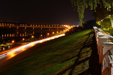 Night city panorama with brightly lit bridge over river and long exposure car light trails. Urban landscape, long exposure photography.