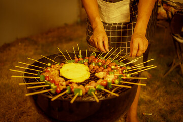 An Asian man wearing a checkered apron carefully arranged skewered meats and vegetables on a round kettle grill. Warm golden lighting highlights the cozy backyard nighttime BBQ atmosphere