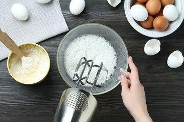 Woman whisking egg whites with hand mixer at black wooden table, top view