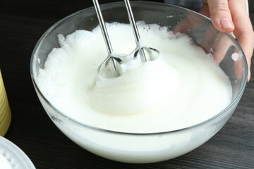 Woman whisking egg whites with hand mixer at black wooden table, closeup