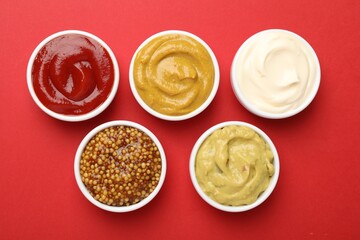 Different sauces in bowls on red background, flat lay