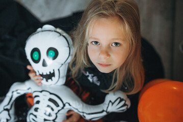 Cute girl in Halloween costume holding inflatable skeleton toy.