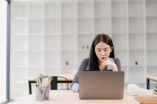 Asian business woman concentrating while working on laptop, sitting at desk in bright modern office, planning or analyzing data, showing professionalism and dedication