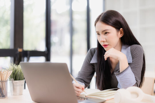 Young Asian woman focusing on work or online learning, sitting at her desk, looking at a laptop computer while holding a pencil and writing in a notebook